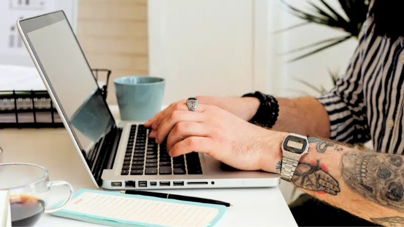 A person with tattooed arms streaming content on a laptop at a desk with a notebook, coffee mug, and plant nearby.