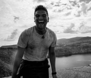 A man wearing a T-shirt and necklace smiles broadly with an open mouth, standing unfiltered in front of a scenic landscape with a body of water and hills in the background.