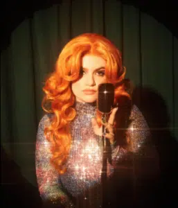 A person with long, wavy orange hair sings into a vintage microphone. They are wearing a sparkling outfit and are illuminated by stage lights.