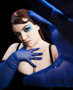 Person with blue eye makeup and blue gloves posing with one hand on their chest and the other extended towards the camera against a black background.