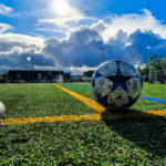 Soccer ball centered on a synthetic field with goalposts in the background, under a cloudy sky. foreground shows a partial view of a soccer shoe.