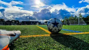 Soccer ball centered on a synthetic field with goalposts in the background, under a cloudy sky. foreground shows a partial view of a soccer shoe.