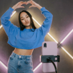 Person in blue sweatshirt and jeans forms a heart shape with their hands while posing in front of a smartphone on a tripod, set against a background with diagonal LED light strips, perfect for their latest TikTok.