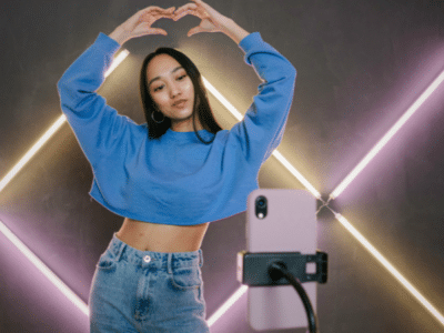 Person in blue sweatshirt and jeans forms a heart shape with their hands while posing in front of a smartphone on a tripod, set against a background with diagonal LED light strips, perfect for their latest TikTok.