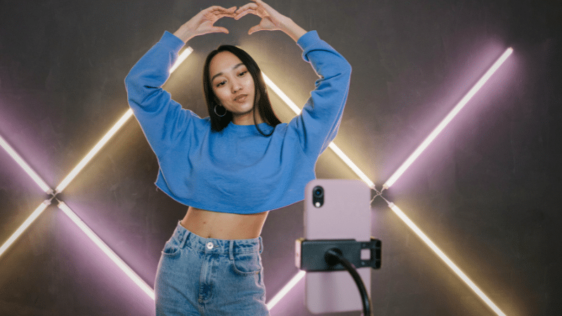 Person in blue sweatshirt and jeans forms a heart shape with their hands while posing in front of a smartphone on a tripod, set against a background with diagonal LED light strips, perfect for their latest TikTok.
