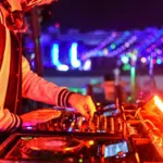 A DJ wearing a jacket and headphones operates a mixing console at a vibrant, brightly lit night event, expertly playlisting tracks to keep the energy high.