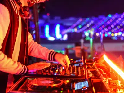 A DJ wearing a jacket and headphones operates a mixing console at a vibrant, brightly lit night event, expertly playlisting tracks to keep the energy high.