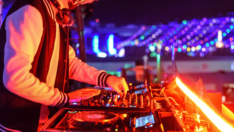 A DJ wearing a jacket and headphones operates a mixing console at a vibrant, brightly lit night event, expertly playlisting tracks to keep the energy high.