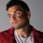 A man with short curly hair, wearing a red jacket over a white shirt, looks to the side with an unfiltered, serious expression. The background is plain and gray.