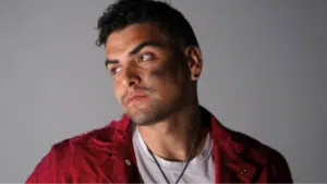 A man with short curly hair, wearing a red jacket over a white shirt, looks to the side with an unfiltered, serious expression. The background is plain and gray.