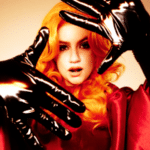 Person with long, bright orange hair wearing a red outfit and shiny, black gloves, holding hands up towards the camera against a beige background.