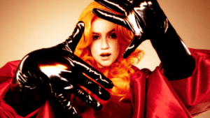 Person with long, bright orange hair wearing a red outfit and shiny, black gloves, holding hands up towards the camera against a beige background.