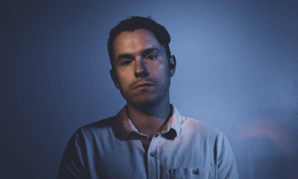 A man with short dark hair is standing against a dark, blue-lit background. He is wearing a light-colored button-up shirt and looking directly at the camera with a neutral expression, perhaps contemplating his latest new music release.