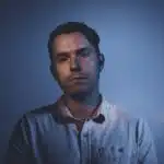 A man with short dark hair is standing against a dark, blue-lit background. He is wearing a light-colored button-up shirt and looking directly at the camera with a neutral expression, perhaps contemplating his latest new music release.