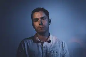 A man with short dark hair is standing against a dark, blue-lit background. He is wearing a light-colored button-up shirt and looking directly at the camera with a neutral expression, perhaps contemplating his latest new music release.