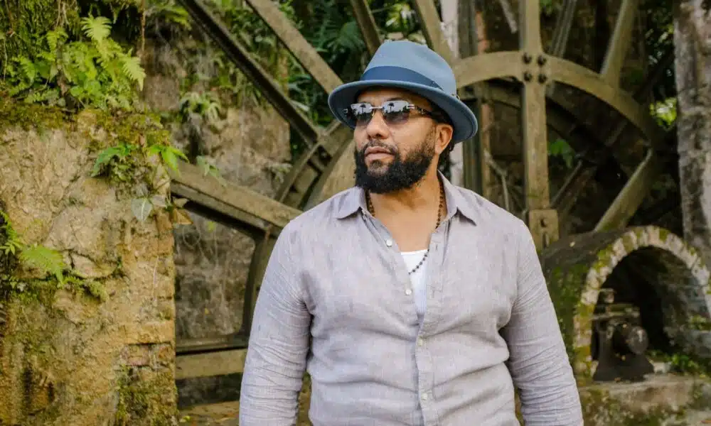 Man with a beard wearing a gray hat and sunglasses standing in front of an old stone water wheel surrounded by lush greenery.