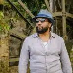 Man with a beard wearing a gray hat and sunglasses standing in front of an old stone water wheel surrounded by lush greenery.