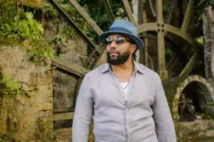 Man with a beard wearing a gray hat and sunglasses standing in front of an old stone water wheel surrounded by lush greenery.