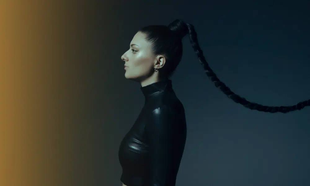 Woman in a side profile view with long braided hair extending horizontally, wearing a black high-neck crop top against a dark background that has a gradient of brown light, ready to unveil her new music.