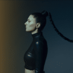Woman in a side profile view with long braided hair extending horizontally, wearing a black high-neck crop top against a dark background that has a gradient of brown light, ready to unveil her new music.