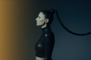 Woman in a side profile view with long braided hair extending horizontally, wearing a black high-neck crop top against a dark background that has a gradient of brown light, ready to unveil her new music.