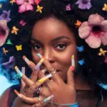 An artist with curly hair adorned with colorful flowers and butterflies, decorated long nails, and rings strikes a pose against a blue background.