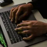 A person is typing on a laptop with green code displayed on the screen in a dimly lit room.