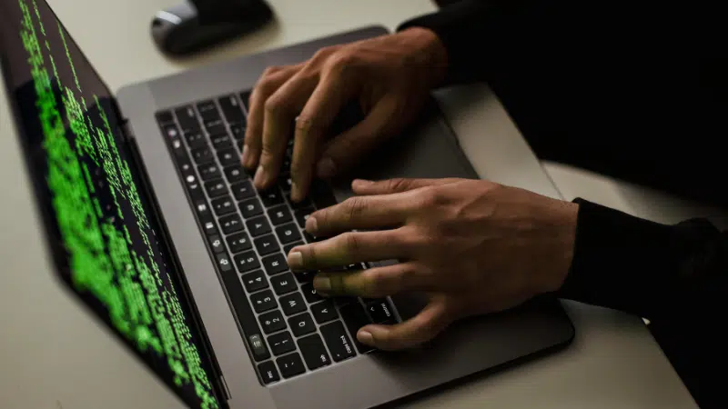 A person is typing on a laptop with green code displayed on the screen in a dimly lit room.