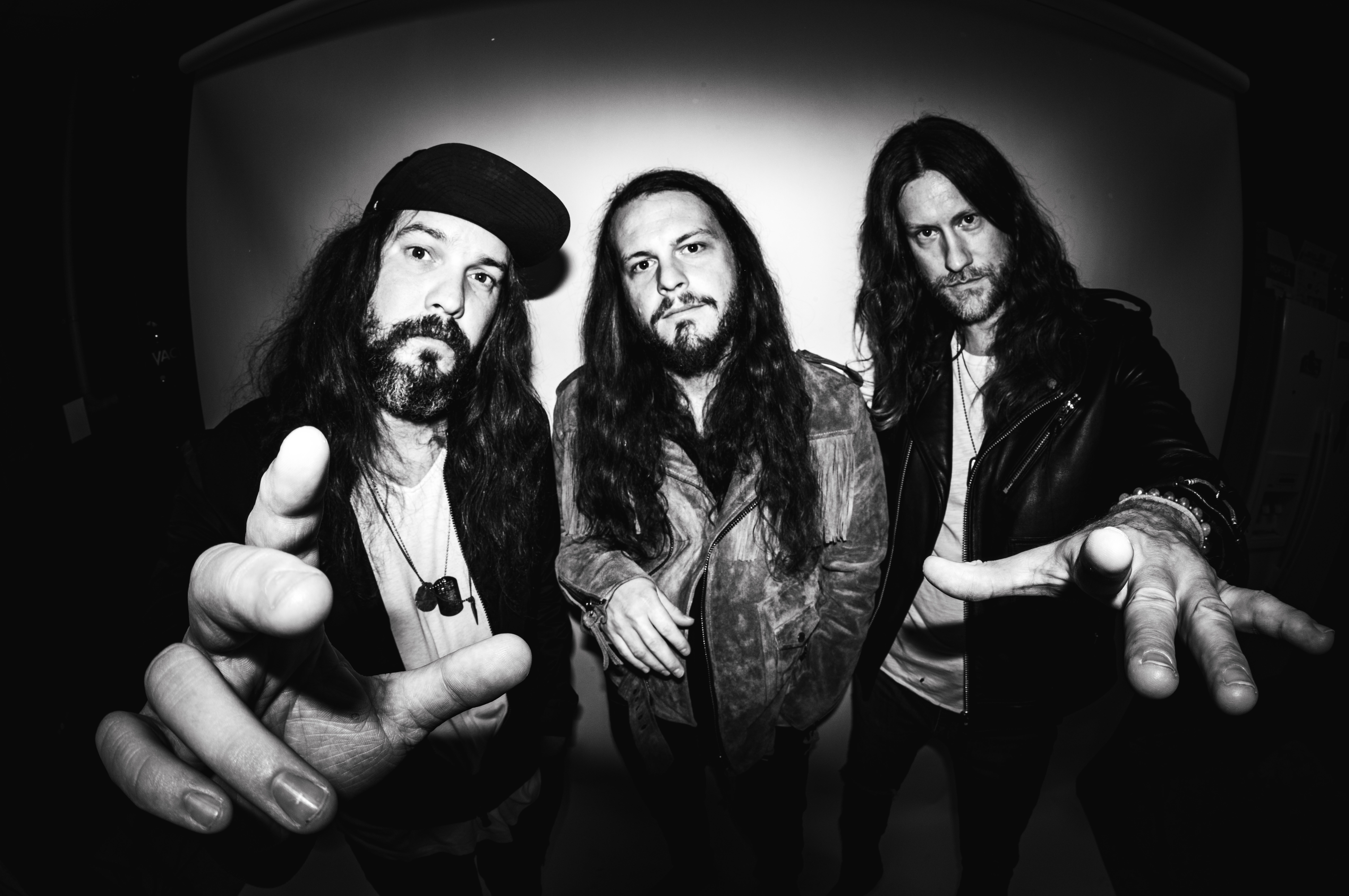 Black-and-white image of three men with long hair, beards, and serious expressions, standing and facing the camera with hands extended towards the lens—could this be a preview of their new music?
