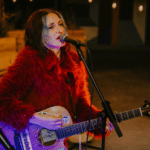 A woman in a red fur coat plays an acoustic guitar and sings into a microphone onstage, with a keyboard in the background, delivering a GRAMMYs-worthy performance.