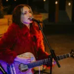 A woman in a red fur coat plays an acoustic guitar and sings into a microphone onstage, with a keyboard in the background, delivering a GRAMMYs-worthy performance.