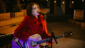 A woman in a red fur coat plays an acoustic guitar and sings into a microphone onstage, with a keyboard in the background, delivering a GRAMMYs-worthy performance.