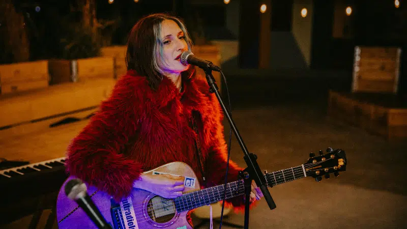 A woman in a red fur coat plays an acoustic guitar and sings into a microphone onstage, with a keyboard in the background, delivering a GRAMMYs-worthy performance.