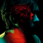 A person with wavy hair is shown in a dark setting with red and green lighting illuminating their face and shoulders.