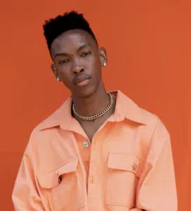 A person wearing an orange shirt and gold chain stands against an orange background, looking into the camera with a neutral expression.