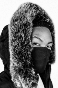 Close-up of a person wearing a fur-trimmed hooded jacket and a black face mask, looking at the camera.