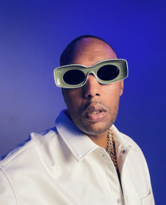 A man wearing angular, futuristic sunglasses and a white jacket poses unfiltered against a blue background.