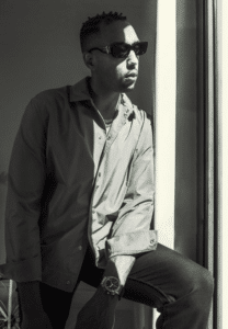 Black and white photo of a man wearing sunglasses and a collared jacket, leaning near a window with unfiltered natural light streaming in.