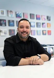 A man with a short beard and mustache, wearing a black shirt, sits at a white table in an office with a wall displaying multiple colorful frames in the background.