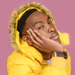 A person in an unfiltered yellow hooded jacket rests their face on their hand against a pink background.