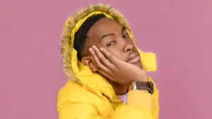 A person in an unfiltered yellow hooded jacket rests their face on their hand against a pink background.