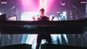 A DJ performs on stage with vibrant pink and purple lights in the background and electronic equipment in front of them.