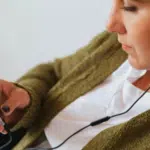 A person wearing headphones looks at a smartphone while leaning back. The individual is wearing a green sweater and white shirt.