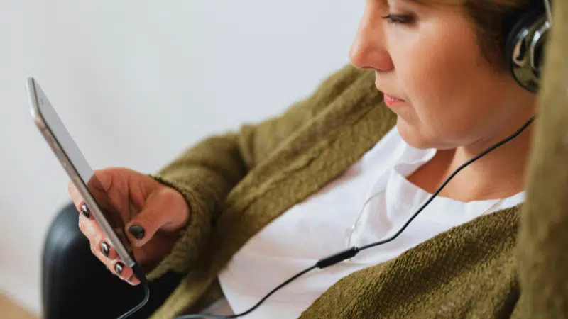 A person wearing headphones looks at a smartphone while leaning back. The individual is wearing a green sweater and white shirt.