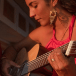 A person wearing a pink top is playing an acoustic guitar with a serious expression, bathed in warm light. The scene evokes the passion of a Latin GRAMMY performance. Another person is partially visible to the left.