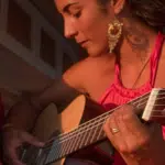 A person wearing a pink top is playing an acoustic guitar with a serious expression, bathed in warm light. The scene evokes the passion of a Latin GRAMMY performance. Another person is partially visible to the left.