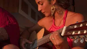 A person wearing a pink top is playing an acoustic guitar with a serious expression, bathed in warm light. The scene evokes the passion of a Latin GRAMMY performance. Another person is partially visible to the left.