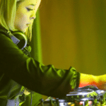 A DJ with headphones around their neck operates mixing equipment, bathed in green and yellow stage lighting, seamlessly curating the night’s vibes straight from their Beatport playlist.