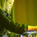 A DJ with headphones around their neck operates mixing equipment, bathed in green and yellow stage lighting, seamlessly curating the night’s vibes straight from their Beatport playlist.