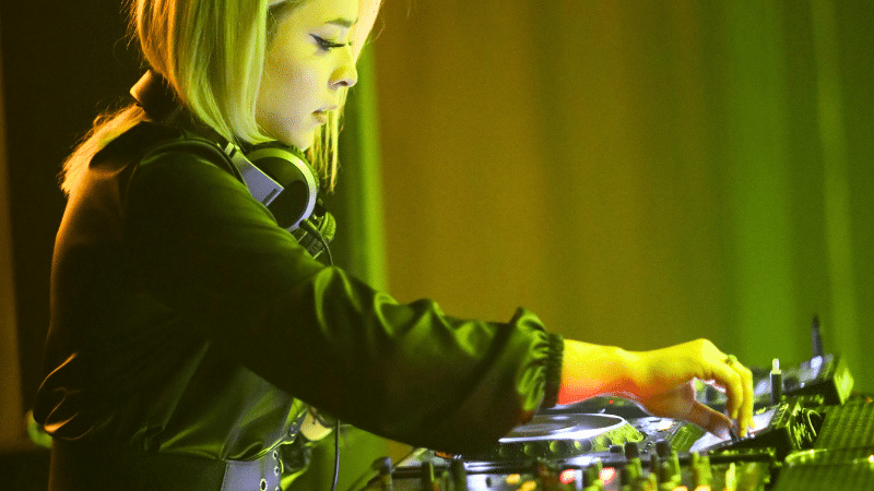 A DJ with headphones around their neck operates mixing equipment, bathed in green and yellow stage lighting, seamlessly curating the night’s vibes straight from their Beatport playlist.